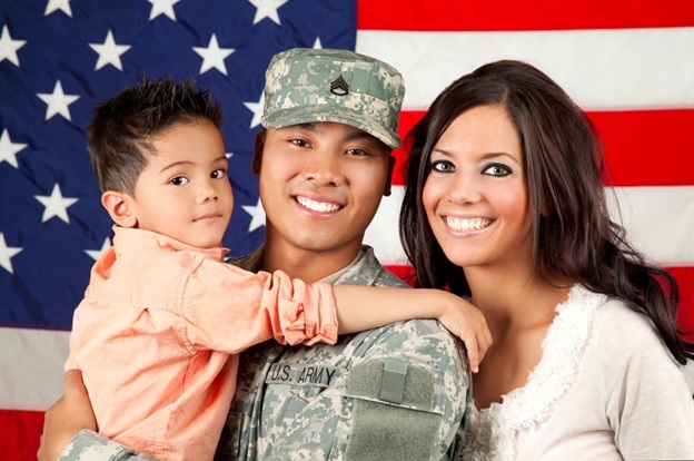 Image of military family with the wife behind the service member who is carrying a young boy in his arms, with a backdrop of the american flag